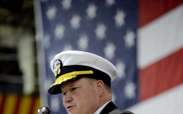 A senior U.S. Navy officer in dress uniform is speaking at a podium with a microphone, in front of a large American flag. The officer displays an extensive array of medals and ribbons on his chest, indicating significant service and commendations. The setting appears to be an official military ceremony, likely held in a hangar bay or large indoor military facility.