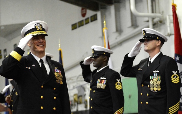 A formal U.S. Navy military ceremony is taking place in what appears to be a ship's hangar bay. Multiple naval officers and enlisted personnel in dress blue uniforms are rendering salutes simultaneously. Large decorative brass shell casings are visible in the foreground, and a red state flag (Arkansas) is displayed to the right.