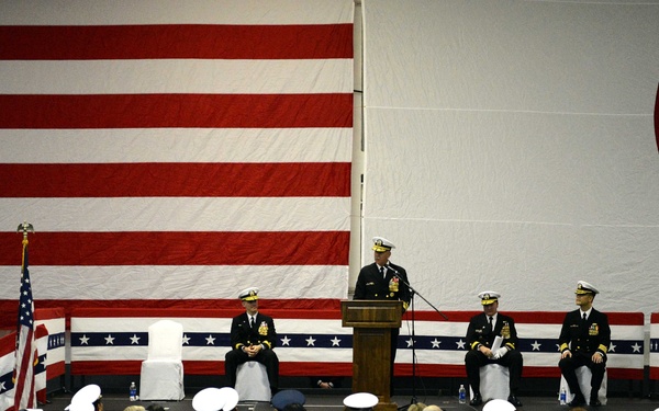 A formal U.S. Navy military ceremony is taking place in an indoor venue, likely a hangar or large hall. A uniformed officer is speaking at a podium while several senior officers are seated on a raised stage. The audience consists of a mix of uniformed Navy personnel and civilians seated in rows of white chairs.