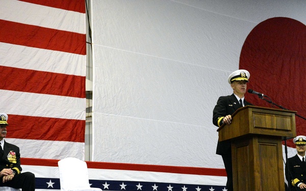 Three U.S. Navy officers in dress uniforms are on a decorated stage during a formal military ceremony. One officer stands at a wooden podium speaking into a microphone, while two others are seated on either side. The stage and backdrop are decorated with large American and Japanese flags, suggesting a bilateral or joint military event.