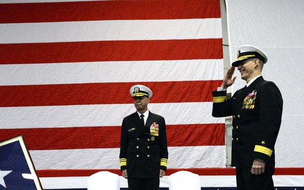 A formal U.S. Navy military ceremony is taking place on a stage in front of a large American flag. Four naval officers in dress blue uniforms are participating, with two exchanging salutes while one holds a framed flag display. The scene appears to be a change of command, retirement, or promotion ceremony based on the formal staging and presentation of a framed flag.