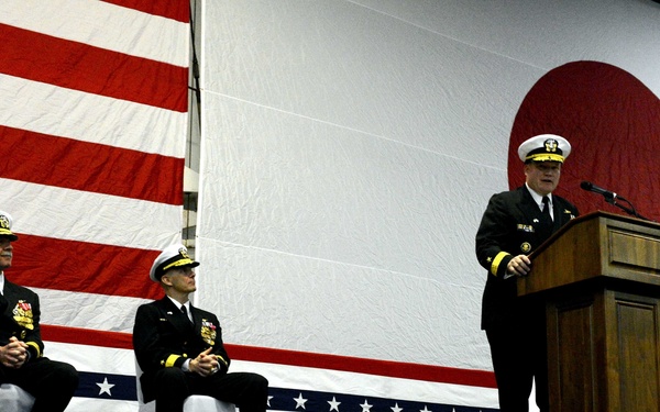 A formal U.S. Navy ceremony is taking place on a decorated stage, with one officer speaking at a wooden podium while two seated officers listen. The stage and backdrop are draped with large American flag bunting, and a large circular red-and-white emblem is visible on the wall behind the speakers. The uniformed personnel are wearing Navy dress blues with gold insignia, suggesting flag or senior officer rank.