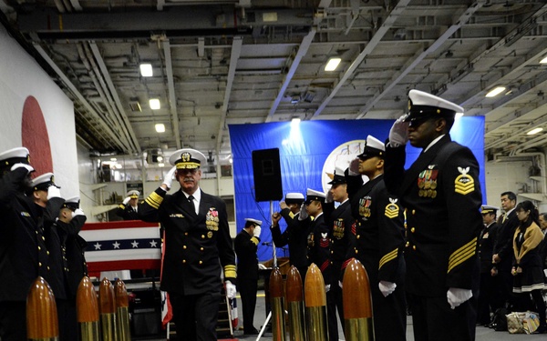 A formal U.S. Navy military ceremony is taking place in what appears to be the hangar bay of an aircraft carrier. A senior naval officer in dress uniform walks down a red carpet flanked by sailors standing at salute, with decorative brass shell casings lining the aisle. A flag-draped casket is visible in the background, suggesting this may be a memorial or funeral ceremony.