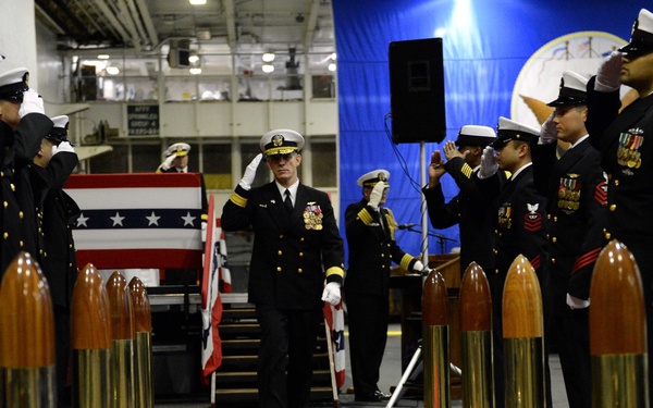 A formal U.S. Navy change of command or retirement ceremony is taking place in what appears to be a ship's hangar bay. A senior naval officer in dress blues walks a red carpet flanked by decorative brass shell casings while personnel on both sides render salutes. An American flag and a blue ceremonial backdrop with a naval insignia are visible in the background.