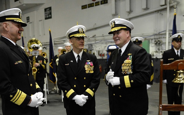 A formal U.S. Navy ceremony taking place in what appears to be a ship's hangar bay. Three senior naval officers in dress uniforms are conversing in the foreground, while a military band and additional personnel are visible in the background. A ship's bell on a wooden stand is visible to the right, indicating this is likely a change of command or commissioning ceremony.