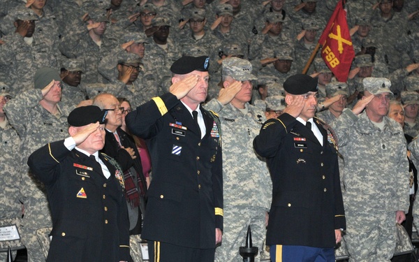 A formal U.S. Army military ceremony, likely a change of command or unit activation, taking place in an indoor gymnasium or arena. Three senior officers in dress blue uniforms stand at the forefront rendering salutes, while a large formation of soldiers in ACU (Army Combat Uniform) camouflage also salute in the background. Unit guidons and flags are visible, indicating an official ceremonial event.