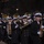National Guardsmen support 57th Presidential Inaugural Parade A military marching band performs during a nighttime parade, with musicians dressed in dark navy uniforms and white sailor caps. The band includes a variety of brass and woodwind instruments, marching in formation along a street lined with spectators. The setting appears to be a formal ceremonial parade, likely in Washington D.C. based on the urban backdrop.