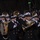 National Guardsmen support 57th Presidential Inaugural Parade A military marching band performs during a nighttime parade, with musicians dressed in dark navy uniforms with gold buttons and white peaked caps. The band includes a variety of brass and woodwind instruments, with a large crowd of spectators visible in the background. The setting appears to be an outdoor urban parade route lined with bare winter trees.