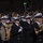 National Guardsmen support 57th Presidential Inaugural Parade A military marching band performs at a nighttime parade before a large crowd of spectators. The musicians are dressed in dark navy uniforms with white peaked caps and white gloves, playing a variety of brass and woodwind instruments. The setting appears to be a formal public event, likely along a major boulevard lined with bare winter trees.