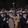 National Guardsmen support 57th Presidential Inaugural Parade A military marching band, identifiable by dark navy uniforms with gold buttons and white peaked caps, performs during a nighttime parade before a large crowd of spectators. The musicians are playing various instruments including clarinets, saxophones, sousaphones, and a bass drum. The setting appears to be an outdoor parade route lined with bare winter trees and street lamps.
