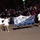 Majestic Marching Knights pass presidential review booth The Ballou High School Majestic Knights Marching Band from Washington, D.C. is performing in a nighttime parade before a large crowd. Band members are dressed in distinctive white jackets and gold pants with white boots, carrying a blue and gold banner identifying the group. The event appears to be a major public parade, likely an inaugural or national celebration given the patriotic bunting visible in the background.