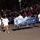Majestic Marching Knights pass presidential review booth The Ballou H.S. Majestic Knights Marching Band from Washington, D.C. is performing in a nighttime parade before a large crowd. Band members are dressed in white military-style uniforms with gold pants and white boots, carrying a large blue and gold banner. The event appears to be a major public parade, likely an inaugural or holiday parade given the patriotic bunting decorations visible on the barriers.
