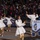 Majestic Marching Knights pass presidential review booth Members of the Ballou High School Marching Band ('Knights') from Washington, D.C. are performing in an outdoor parade at night. The performers, dressed in white coats and gold metallic pants with gold gloves, are dancing and marching in formation. A large crowd of spectators lines the parade route behind metal barricades.