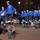 Gym Dandies dazzle crowd at 57th Presidential Inauguration Parade A large group of performers dressed in matching blue jackets and black pants are performing a unicycle routine during what appears to be a parade or public event at night. One performer stands out by riding a tall stacked unicycle with three wheels (red, white, and blue colored) while juggling. The crowd of spectators lines the barriers in the background.
