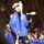 Gym Dandies dazzle crowd at 57th Presidential Inauguration Parade A performer in a blue jacket and white bicycle helmet is juggling white clubs during what appears to be a nighttime parade or outdoor performance. Multiple other performers in matching blue uniforms and white helmets are visible in the background, along with a crowd of spectators. The setting appears to be along a tree-lined street or path illuminated by artificial lighting.
