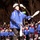 Gym Dandies dazzle crowd at 57th Presidential Inauguration Parade A performer in a blue jacket and white helmet is juggling white clubs during what appears to be a parade or public performance event. Behind the lead performer, a large group of similarly uniformed performers wearing matching blue jackets, helmets, and knee pads are visible. The event appears to take place at night or in low-light conditions along a street lined with bare winter trees and a large crowd of spectators.