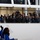 Gym Dandies dazzle crowd at 57th Presidential Inauguration Parade A group of spectators watches performers in blue uniforms labeled 'Gym Dandies' riding unicycles in what appears to be an indoor venue with a glass-enclosed viewing area. The crowd in the elevated viewing area includes formally dressed individuals, some with cameras, observing the unicycle performance below. The setting appears to be an official or ceremonial event given the formal attire and presence of what appears to be media personnel with cameras.