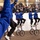 Gym Dandies dazzle crowd at 57th Presidential Inauguration Parade A group of performers in blue jackets and white helmets are riding unicycles in what appears to be an indoor parade or ceremonial performance. They are performing before a crowd of spectators watching from behind a glass barrier in what looks like an official viewing stand. The performers are waving to the audience while maintaining balance on their unicycles.