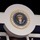 National Guardsmen support 57th Presidential Inauguration The image shows a large decorative display of the official Seal of the President of the United States, mounted on a white circular panel above what appears to be a booth or stage structure. The seal features the traditional bald eagle design with a shield, olive branch, and arrows, surrounded by stars and the official inscription. The setting appears to be an indoor venue or exhibition hall with recessed lighting visible in the background.