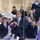 National Guardsmen support 57th Presidential Inaugural Parade A formal public event, likely a parade or inauguration ceremony, showing a group of well-dressed individuals in what appears to be a viewing stand or enclosed booth. A central figure in a dark suit and blue tie is standing and applauding with a broad smile, surrounded by other formally dressed attendees. Purple streamers or ribbons are visible in the foreground, suggesting a celebratory occasion.