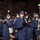 United States Air Force Band passes presidential reviewing stand A military marching band performs during a nighttime parade, with members dressed in dark navy blue uniforms and matching caps. The band includes a variety of instruments including clarinets, saxophones, flutes, and snare drums. A large crowd of spectators is visible in the background along a tree-lined street illuminated by street lamps.