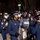 United States Air Force Band passes presidential reviewing stand A military marching band performs during a nighttime parade, with members dressed in navy blue uniforms and matching caps with insignia. The central figure plays a saxophone while surrounded by bandmates playing sousaphones, drums, and other brass instruments. A large crowd of spectators lines the parade route in the background amid bare winter trees.