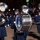 United States Air Force Band passes presidential reviewing stand The United States Air Force Band is performing in a nighttime parade in Washington, D.C. Band members dressed in dark navy blue uniforms and black caps are marching while playing sousaphones and a bass drum. A large crowd of spectators is visible in bleachers in the background.