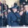 United States Air Force Band passes presidential reviewing stand A formal outdoor public event, likely a government or military ceremony, taking place in cold weather. Three prominent figures stand in the foreground on what appears to be a viewing platform or stage, separated from a crowd by a glass barrier. The central military figure is in uniform, flanked by two men in dark suits with blue ties.