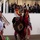 National Guardsmen support 57th Presidential Inaugural Parade Native American dancers in traditional regalia perform at what appears to be a formal official event, with a large audience watching from an elevated viewing area behind glass. The dancers wear elaborate feathered headdresses, colorful beadwork, and traditional regalia including large circular feather bustles. The setting appears to be an indoor parade or ceremonial performance venue.