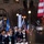 New York National Guardsmen support 57th Presidential Inauguriation A marching band parade is taking place at night in what appears to be New York City, as indicated by a visible 'NEW YORK' banner/sign. Band members are dressed in dark navy blue uniforms with white gloves and hats, carrying instruments including a sousaphone/tuba and flutes. Multiple flags are visible including the American flag, a POW/MIA flag, and New York state flags.