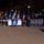 New York National Guardsmen support 57th Presidential Inauguriation A nighttime parade featuring a New York delegation marching with flags, banners, and a state sign. The group carries multiple flags including American flags and what appears to be military/organizational flags, along with a banner reading 'Producers of the New York City Veterans Day Parade.' Crowds of spectators line the barricaded parade route in cold weather attire.