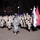 High School JROTC marches in 57th Presidential Inaugural Parade A military parade is taking place at night on a city street, featuring a large formation of uniformed personnel in desert camouflage (DCU) uniforms marching in formation. A single figure in Air Force digital camouflage (ABU pattern) stands facing the formation, appearing to be in a supervisory or command role. Flags including what appears to be a U.S. Marine Corps flag and a blue service flag are being carried by color guard members.