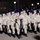 High School JROTC marches in 57th Presidential Inaugural Parade A formation of military personnel or cadets in desert camouflage (DCU - Desert Camouflage Uniform) marching in what appears to be a nighttime parade. The participants wear matching tan/brown camouflage uniforms, black gloves, and patrol caps, and carry what appear to be rifles or flagpoles. A large crowd of spectators is visible in the background along a parade route.