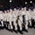 High School JROTC marches in 57th Presidential Inaugural Parade A formation of military personnel or cadets in desert camouflage (DCU - Desert Combat Uniform) marching in what appears to be a nighttime parade. One individual in the front carries a dark pennant or guidon bearing the text 'OK-20012'. Spectators and bleachers are visible in the background, suggesting a formal ceremonial event.