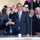 High School JROTC marches in 57th Presidential Inaugural Parade A man in a dark navy suit with a light blue tie stands at the center of the image, smiling broadly and waving with his right hand raised. He is surrounded by a crowd of people, some in formal attire, behind what appears to be a transparent protective barrier. A white paper cup is visible on the ledge in front of him.