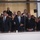 National Guardsmen support 57th Presidential Inaugural Parade A group of formally dressed individuals stand behind a glass barrier, with a central figure in a dark suit and light blue tie waving to a crowd. The setting appears to be an official or ceremonial event, possibly a parade reviewing stand or inaugural platform. Multiple people in the background are photographing the scene with phones and cameras.