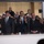 National Guardsmen support 57th Presidential Inaugural Parade A group of formally dressed individuals stand behind a glass partition, likely in a viewing box or enclosed observation area at a public event. The central figure is smiling broadly and clapping, flanked by others in suits. The setting suggests an official or ceremonial occasion, with a crowd visible in the background.