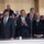 National Guardsmen support 57th Presidential Inaugural Parade A group of formally dressed individuals stand behind a glass barrier, with a smiling man in the center clapping his hands. A Presidential seal cup is visible on the ledge in front of them. The setting appears to be an official or ceremonial event with a crowd visible in the background.