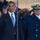 President Obama salutes the United States Coast Guard The image shows two individuals in the foreground rendering a salute at what appears to be a formal ceremonial event. One person is wearing a dark business suit with a blue tie and an American flag lapel pin, while the other is dressed in a dark naval/coast guard dress uniform with a white officer's cap and gold insignia. A crowd of spectators is visible in the background.