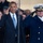 President Obama salutes the United States Coast Guard Two men are rendering a salute in what appears to be a formal outdoor ceremony. The man on the left wears a dark business suit with a blue tie and an American flag lapel pin, while the man on the right wears a dark naval/coast guard dress uniform with gold buttons, black gloves, and a white officer's cap with gold insignia. A crowd of spectators is visible in the background.
