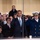 President Obama salutes the United States Coast Guard The image shows a formal ceremonial event where a man in a dark suit with a blue tie is rendering a salute alongside a senior military officer in a dark navy dress uniform with a white peaked cap. They appear to be standing at a reviewing stand, likely during a parade or official ceremony. A crowd of attendees is visible in the background behind a glass partition.