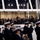 United States Coast Guard marches in 57th Inaugural Parade A military marching band in dark navy uniforms with white caps performs in the foreground, playing brass instruments including tubas and trombones. Behind a glass barrier on an elevated platform, a crowd of dignitaries and spectators watches the performance, including several individuals in military dress uniforms. The setting appears to be an official ceremonial event held at a modern indoor/covered venue.