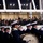 United States Coast Guard marches in 57th Inaugural Parade A military marching band in dark blue uniforms and white caps performs in the foreground, while a crowd of dignitaries and officials watches from an enclosed glass viewing platform above. The setting appears to be a formal ceremonial event, likely an inaugural parade or official state occasion, given the presence of uniformed military officers and formally dressed civilians in the viewing stand. The scene is illuminated by overhead lighting against a dark exterior backdrop.