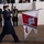 United States Coast Guard marches in 57th Inaugural Parade A military color guard ceremony is taking place, with a cadet in a dark blue dress uniform and white gloves carrying a pennant flag for the United States Coast Guard Academy. Multiple service members from different branches are visible in the background, standing at attention. The setting appears to be an official parade or ceremonial event, likely at a formal venue.
