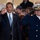 President Obama salutes the United States Coast Guard Two men are saluting in what appears to be a formal outdoor ceremony. One man is wearing a dark business suit with a blue tie and an American flag lapel pin, while the other is dressed in a dark navy military dress uniform with gold buttons and a white officer's cap. A crowd of onlookers is visible in the background.