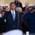 President Obama salutes the United States Coast Guard Two men are saluting in what appears to be an official ceremonial setting. The man on the left wears a dark civilian suit with a blue tie and an American flag lapel pin, while the man on the right wears a dark blue military dress uniform with gold buttons, rank insignia, and a white officer's cap. A large crowd of observers is visible in the background.