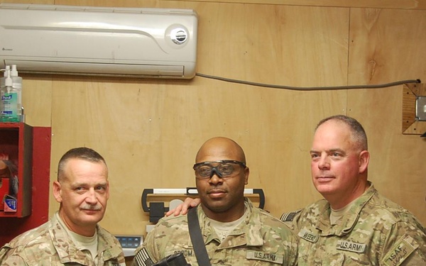 Three U.S. Army soldiers in MultiCam/OCP camouflage uniforms pose together inside what appears to be a forward operating base (FOB) common room or break room. The center soldier is wearing tactical eyewear and carrying a slung rifle. Visible patches and insignia suggest a deployment context, with an ISAF patch visible on the right soldier's uniform.
