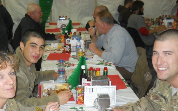 Military personnel in U.S. Army/ISAF camouflage uniforms are gathered for a holiday meal, likely Christmas, in what appears to be a large dining tent or facility. The tables are decorated with Christmas-themed tablecloths featuring holly and poinsettia patterns, along with small green Christmas tree decorations. Three soldiers in the foreground are smiling at the camera while eating from compartmentalized meal trays.