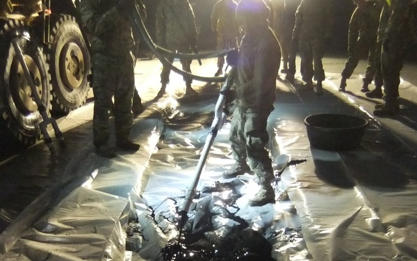 A nighttime scene showing multiple personnel in military-style uniforms working around a large plastic containment sheet or tarp covered with a dark liquid, likely oil or fuel. One individual is using a long-handled tool to manage the liquid on the tarp. A military or heavy-duty vehicle is visible on the left side of the frame.