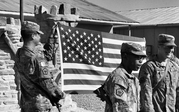 A black-and-white photograph showing U.S. Army soldiers in MultiCam/OCP camouflage uniforms during what appears to be a flag ceremony at a forward operating base or military installation. At least four soldiers are visible, with one raising or handling a large American flag attached to a flagpole near a stone structure. The setting includes gravel ground, stone masonry walls, and prefabricated military-style buildings in the background.